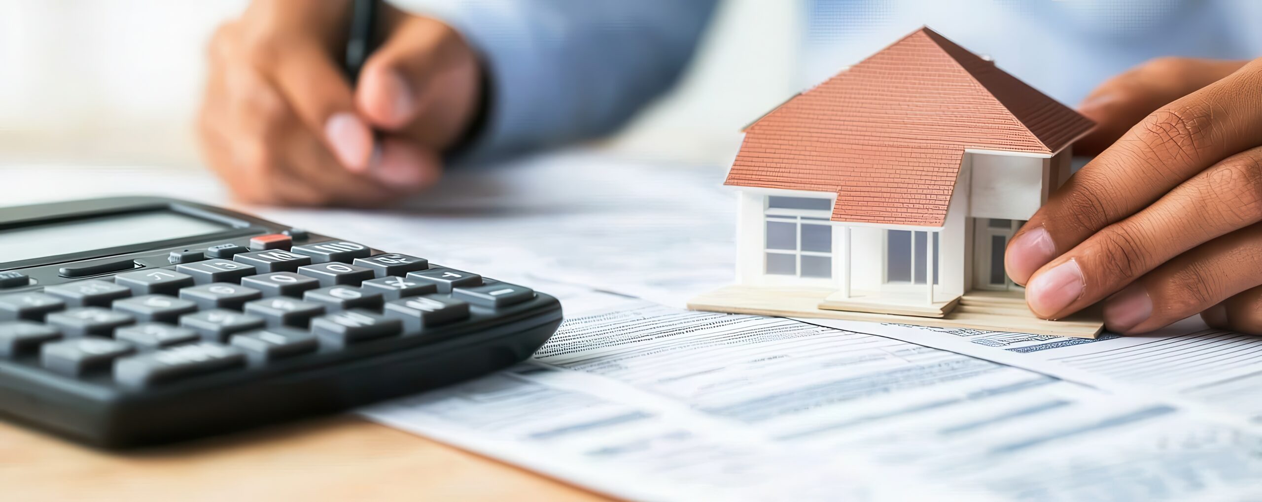 A person’s hand holds a miniature house model while the other hand writes notes next to a calculator and paperwork, symbolizing real estate transactions.